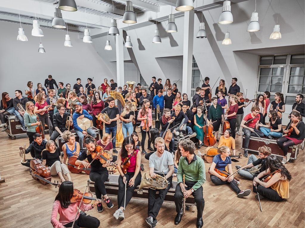 Die vielen Mitglieder des Orchesters stehen und sitzen in bunter Kleidung und mit ihren Instrumenten in einem großen Raum auf Kisten und dem Boden. Sie lachen, sprechen miteinander oder spielen auf ihrem Instrument.