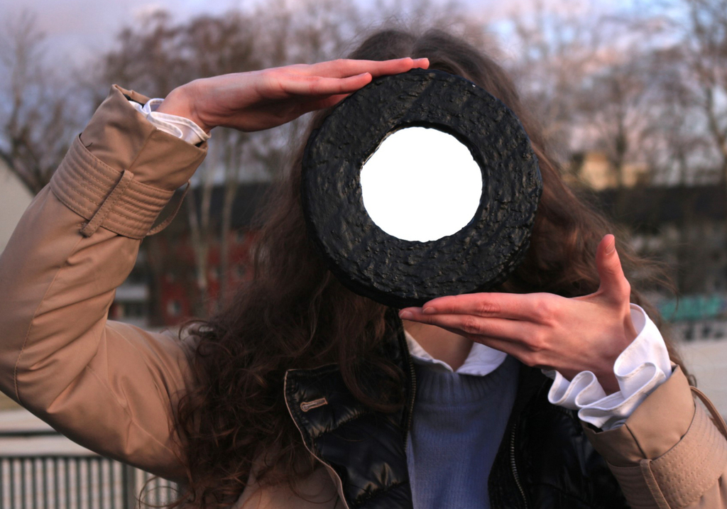 Eine brünette Frau steht in herbstlicher Kleidung auf einem Platz und hält einen Spiegel vor ihr Gesicvht, sodass man den bedeckten Himmel darin sieht