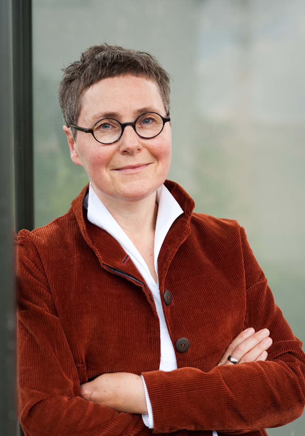 Porträt der Autorin in rostroter Jacke, angelehnt an einen Türrahmen