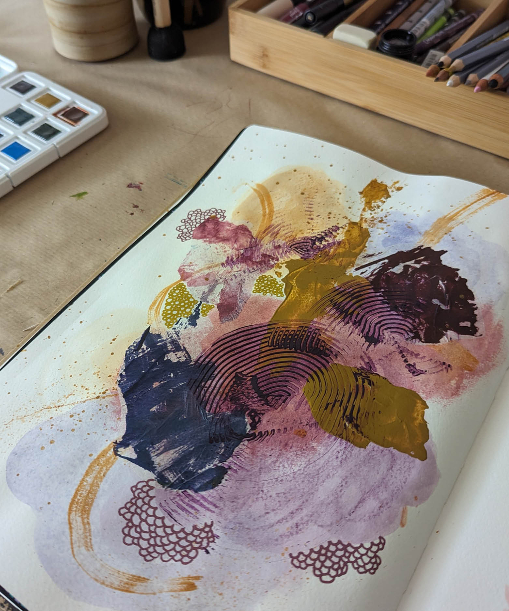 Ein aufgeschlagenes Notizbuch mit einem bunt bemalten Blatt, im Hintergrund verschiedene Stifte und ein Farbkasten