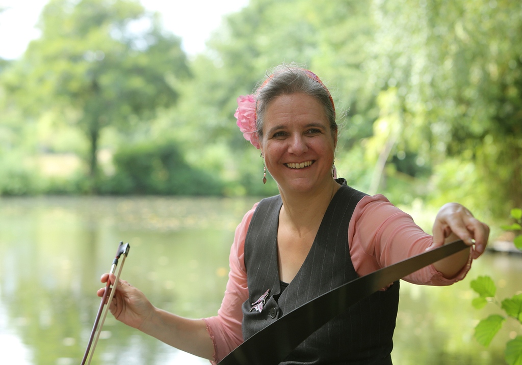 Ein Foto von Susanne Sewald mit ihrem Instrument, lachend, mit Blume im Haar und an einem See sitzend