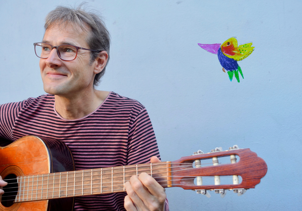 Der Musiker Jens Digel sitzt mit seiner Gitarre vor einem blauen Hintergrund, oben rechts ist ein bunter, selbstgemalter Vogel  