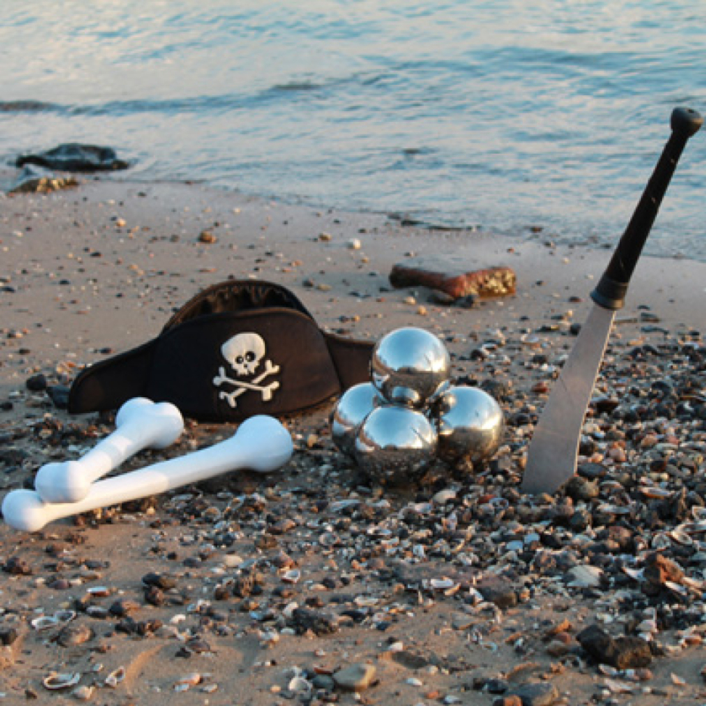 Ein Piratenhut, Plastikknochen, Kanonenbälle aus Metall und eine Messerattrape stecken im Sand eines Strandes, im Hintegrund das Meer