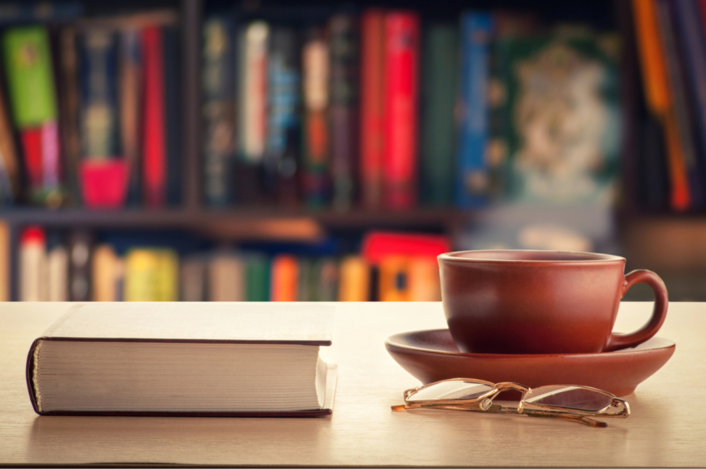 Eine dampfende Tasse Tee oder Kaffee steht auf einem Tisch neben einem Stapel Bücher. Im Vordergrund liegt eine Brille, im Hintergrund sind Bücherregale zu sehen