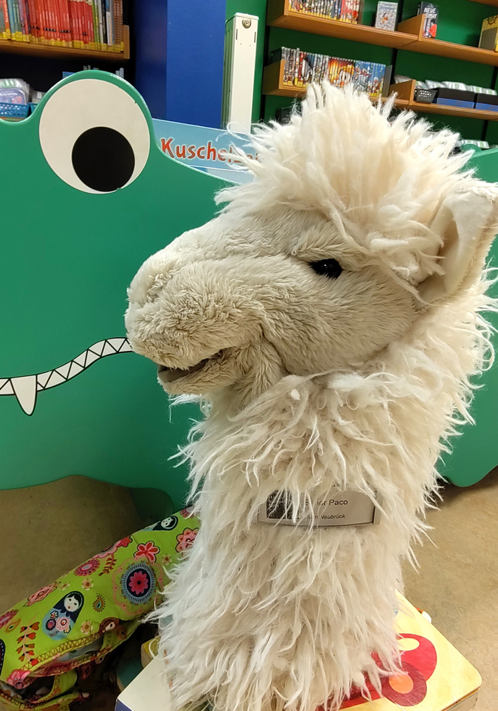 Ein Stoff-Alpaca mit Namensschild "Senor Paco" sitzt auf einem Bücherstabel vor einem Bilderbuchtrog in Krokodilform