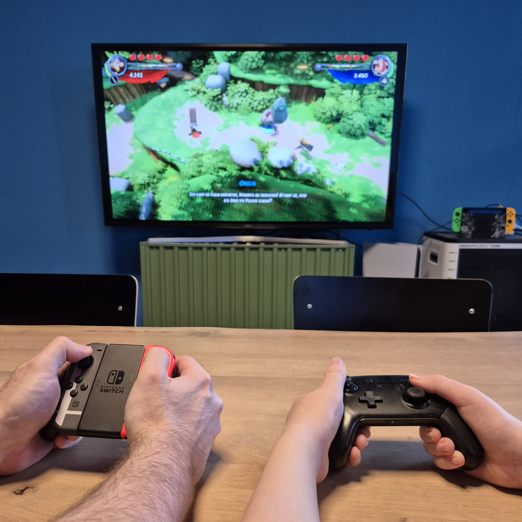 Die Hände von zwei Personen an der Spieleconsole mit Blick auf einen Fernseher, an dem ein Computerspiel läuft