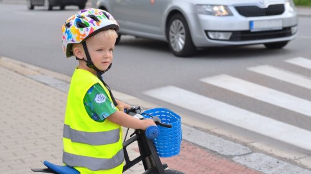 Kleiner Junge mit Warnweste, Fahrradhelm und  Laufrad wartet an einem Zebrastreifen. Ein Auto hält vor dem Zebrastreifen.