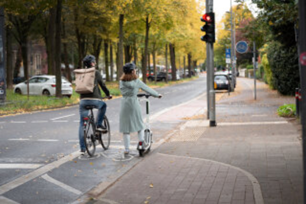 Zu sehen sind ein Mann auf einem Fahrrad und eine Frau auf einem E-Scooter, die nebeneinander an einer roten Ampel warten. Das Rotsignal der Ampel ist in Herzform., Link auf Bleib stehen bei Rot – jemand liebt dich!