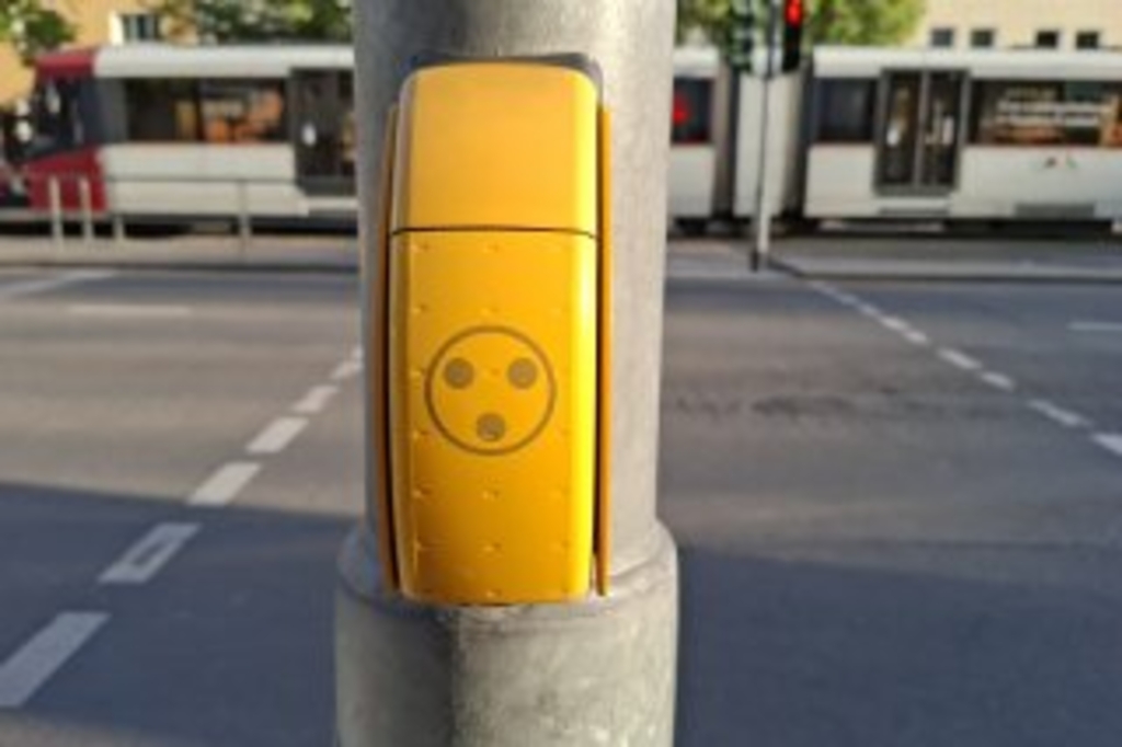 Zu sehen ist ein Taster an einem Ampelmast mit einem Symbol mit drei schwarzen Punkten in einem schwarzen Kreis.