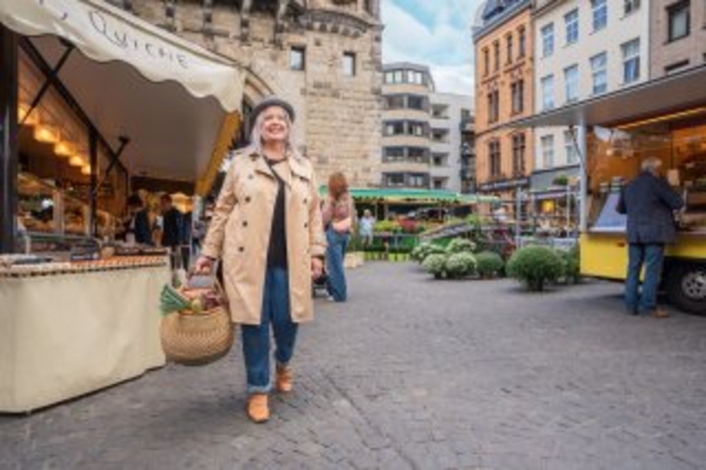 Plakatmotiv der Aktion "Stadt, Land, zu Fuß". Eine Frau mit Hut, Mantel und Einkaufskorb voller Gemüse geht über einen Kölner Wochenmarkt., Link auf Stadt, Land, zu Fuß