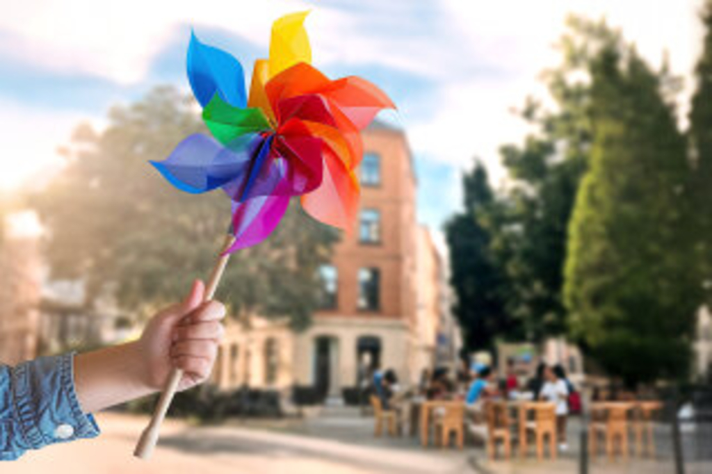 Windrädchen in Kinderhand. Im Hintergrund sieht man die Fassade eines Hauses mit Menschen auf Stühlen davor., Link auf Bürgerrat "Mobil im lebenswerten Quartier"