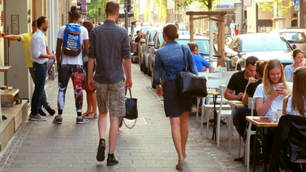 Gehweg, auf dem mehrere Menschen gehen. Rechts sind Tische, an denen Menschen sitzen., Link auf Gestaltungshandbuch