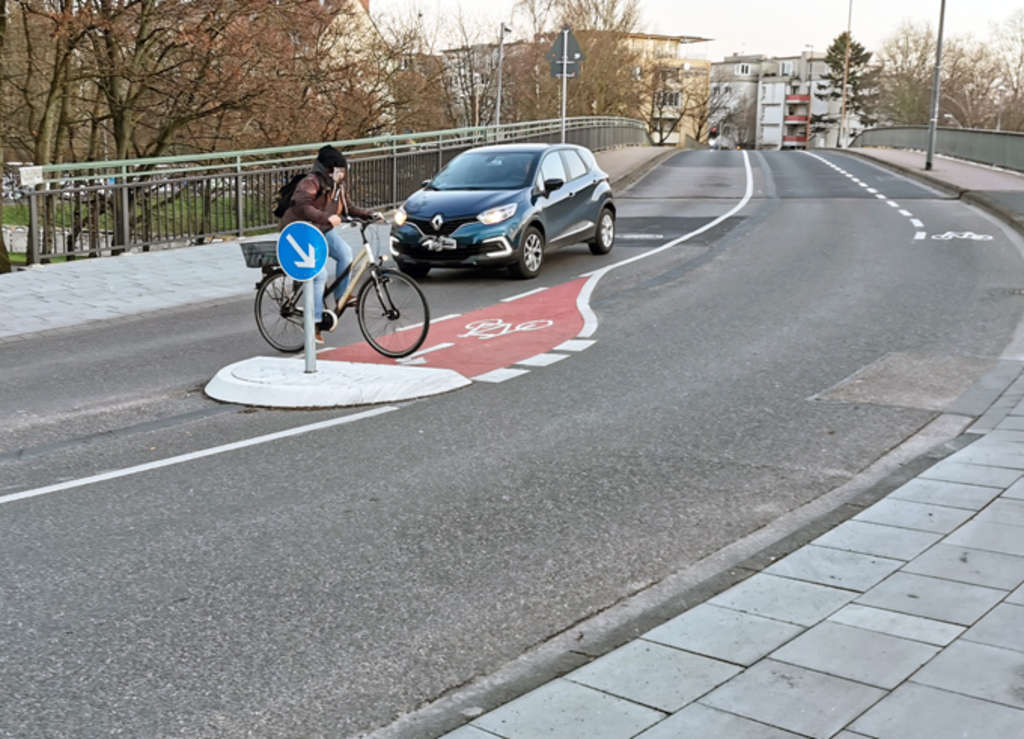 Radfahrer überquert die Straße von links nach rechts hinter einer Brücke. 
