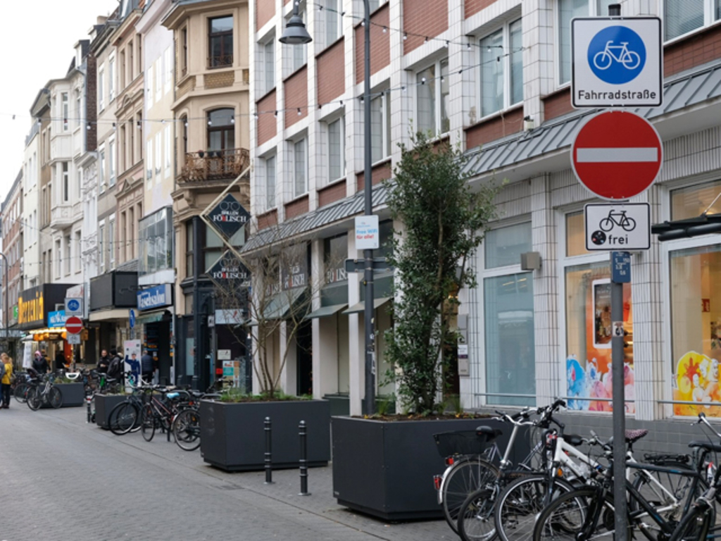 Blick auf den Eigelstein nach der Verkehrsberuhigung und städtebaulichen Aufwertung. Zu sehen sind auf der rechten Seite Baumkübel, ein Fahrradfahrer fährt den Eigelstein entlang. Viele Passant*innen sind unterwegs.