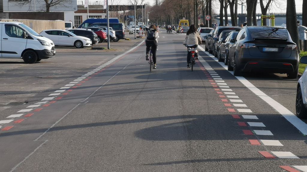 Zwei Radfahrerinnen fahren über die neugestaltete Alfred-Schütte-Allee. Zu sehen sind Parkstände und die neue rote Fahrradstraßenlinie.