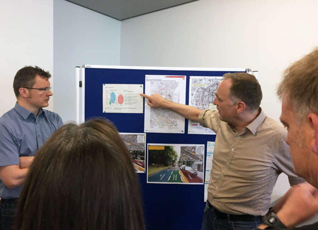 Zwei Mitarbeiter der Stadt Köln erklären etwas vor einer Stellwand