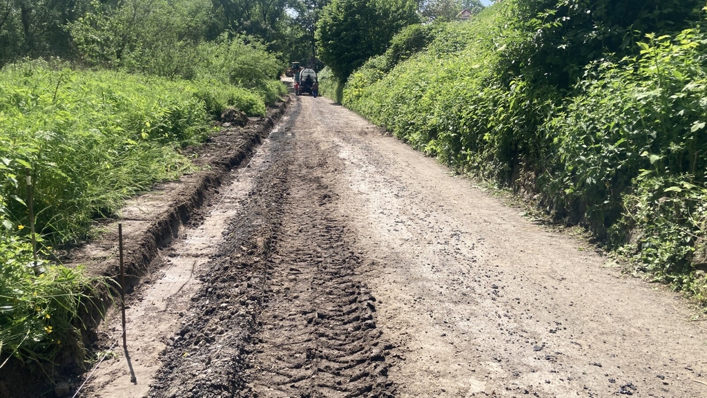 Auf dem angehängten Foto sehen Sie einen Abschnitt des in Bau befindlichen Geh- und Radweg am Sürther Leinpfad. Aktuell wird dort der Untergrund vorbereitet und der zukünftige Weg eingemessen. Links und rechts des Weges befinden sich Grünflächen sowie im hinteren Teil des Bildes verschiedene Baufahrzeuge.