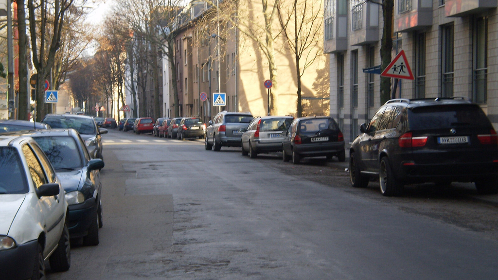 Blick in die Takustraße. Der Straßenbelag ist in einem sanierungsbedürftigen Zustand. An den Straßenseiten stehen geparkte Autos.
