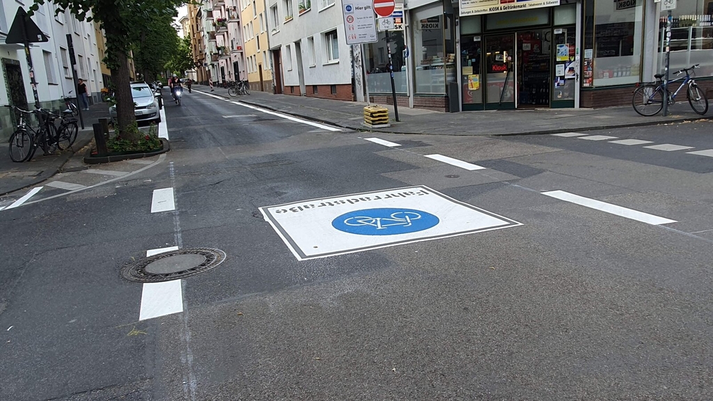 Foto der Fahrradstraße im Pantaleonswall. Zu sehen ist ein großes Fahrradstraßen-Piktrogramm in der Fahrbahnmitte. Autos parken im Seitenbereich. Im Hintergrund sind Radfahrende zu sehen.