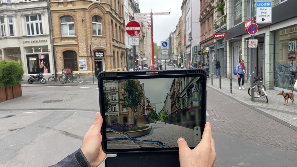 Zu sehen ist ein Foto mit Blick auf der Ehrenstraße. Zu sehen ist ein Tablet, auf dem die neugestaltete Ehrenstraße als Augmented Reality zu sehen ist. Im Hintergrund zu sehen ist die "real" Ehrenstraße. Zu sehen ist, wie die Neugestaltung die Ehrenstraße grüner und schöner macht.