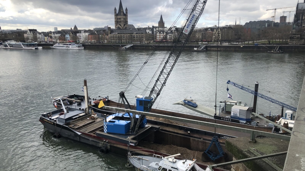 Zu sehen ist ein Bagger, der vom Schiff aus Füllmaterial auf den Rheingrund ablässt.