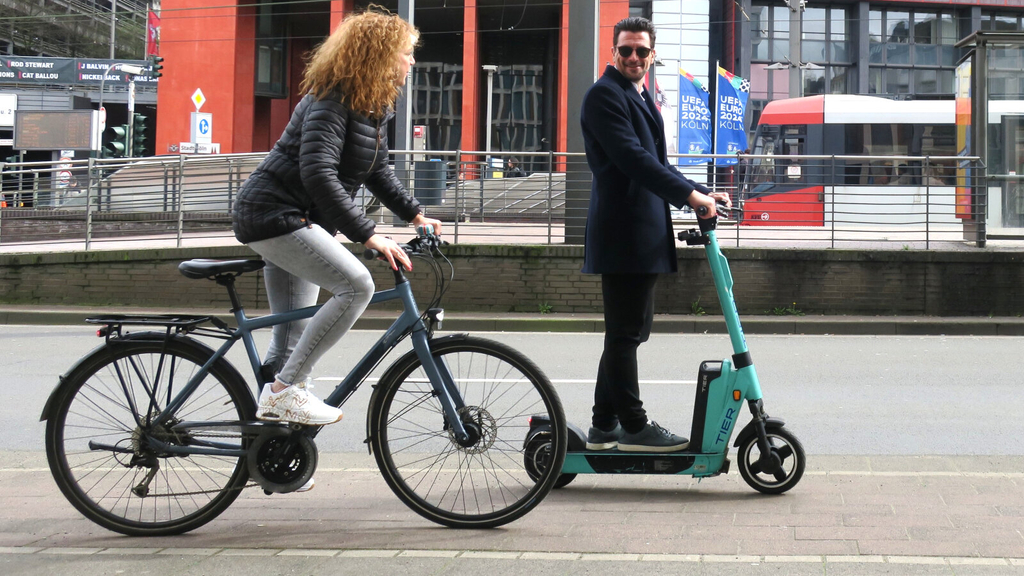 Eine Radfahrende wird auf einem Radweg von einem lächelnden E-Roller-Fahrer überholt.