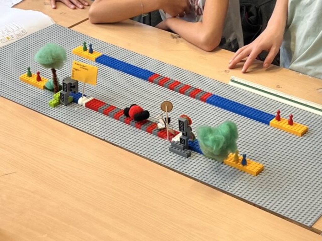 Schüler*innen haben auf einer Legoplatte mit Legosteinen eine Fahrradstraße nachgebaut. Die Schilder sind aus Papier gebastelt. Weiter gibt es selbstgebastelte Blumen und Bäume. Die Menschen sind mit Spielfiguren von Gesellschaftsspielen dargestellt.