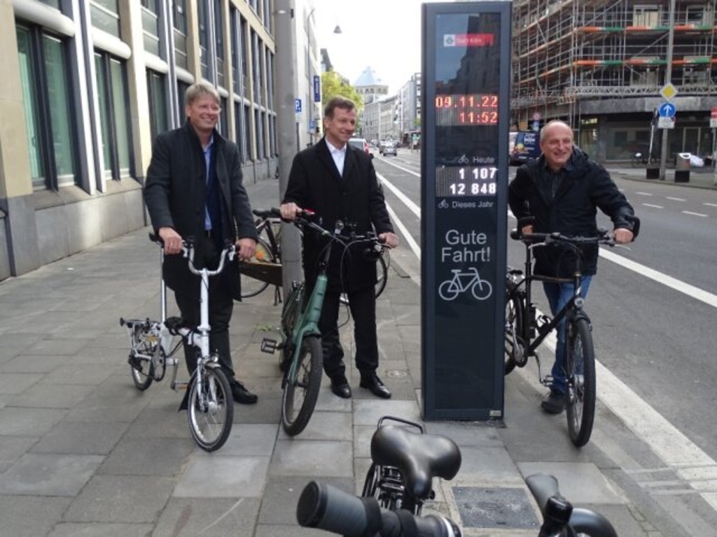 Thorsten Siggelkow, Amtsleiter des Amtes für nachhaltige Mobilität, Ascan Egerer, Beigeordneter für Mobilität und Jürgen Möllers, Fahrradbeauftragter, stehen auf der Gladbacher Straße mit Fahrrad neben dem Fahrradbarometer.