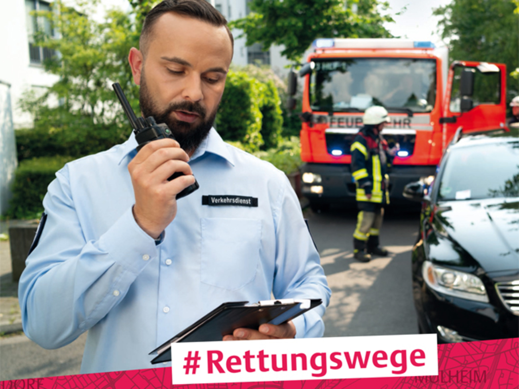 Mitarbeiter des Ordnungsdienstes mit Funkgerät in der Hand, im Hintergrund eine Feuerwehrauto, welches nicht weiterfahren kann, weil ein Fahrzeug die Straße versperrt.