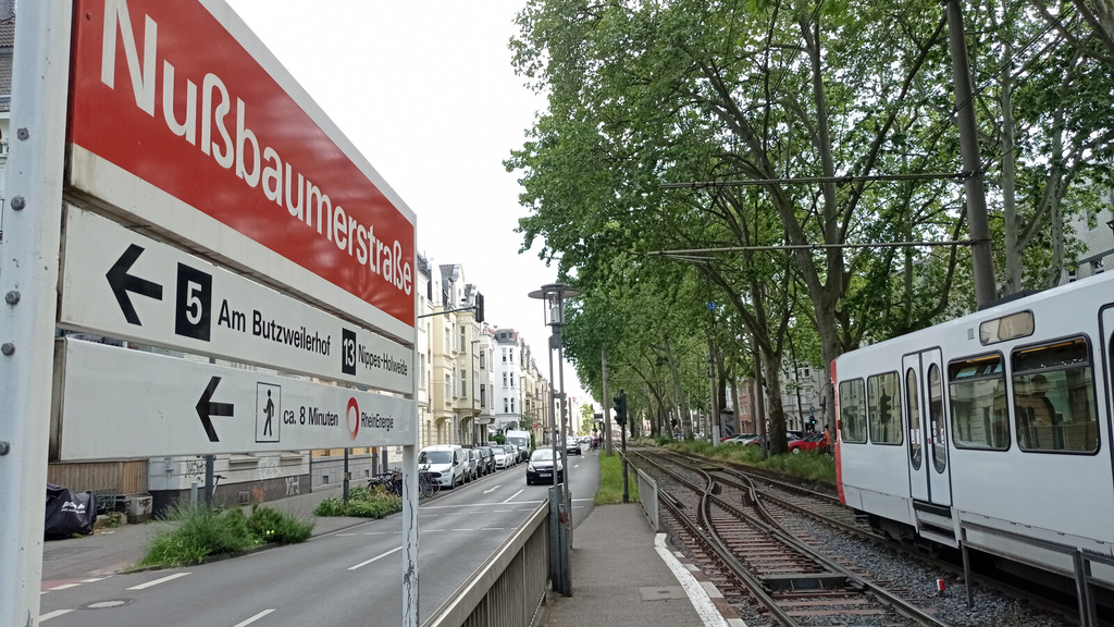 Stadtbahnhaltestelle Nußbaumerstraße. Im Vordergrund ist das Haltestellenschild zu sehen. Seitlich fährt eine Stadtbahn.