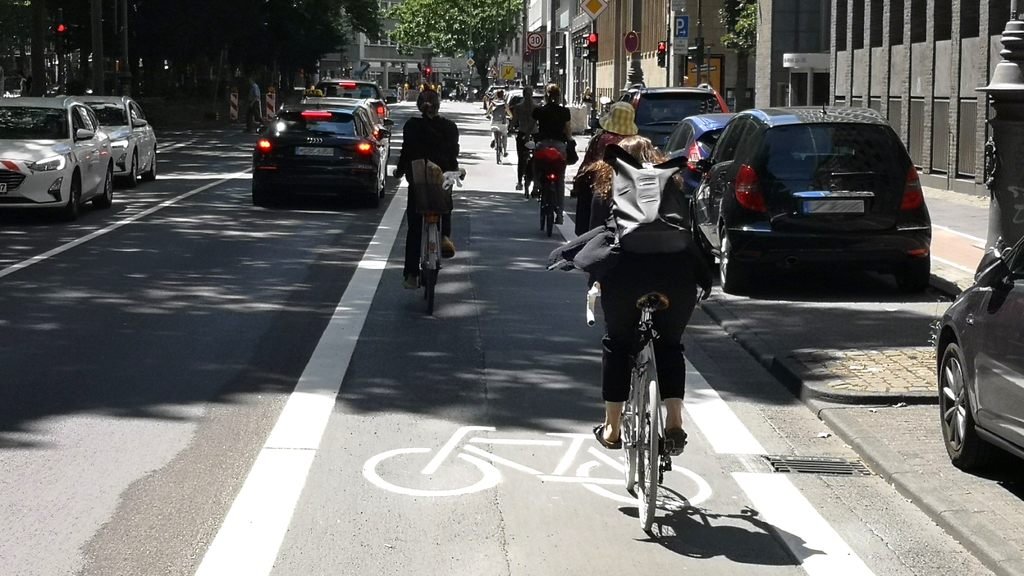 Neuer Radfahrstreifen auf dem Kaiser-Wilhelm-Ring. Zu sehen sind einige Radfahrende, die den Radfahrstreifen befahren sowie parkende und fahrende Autos.