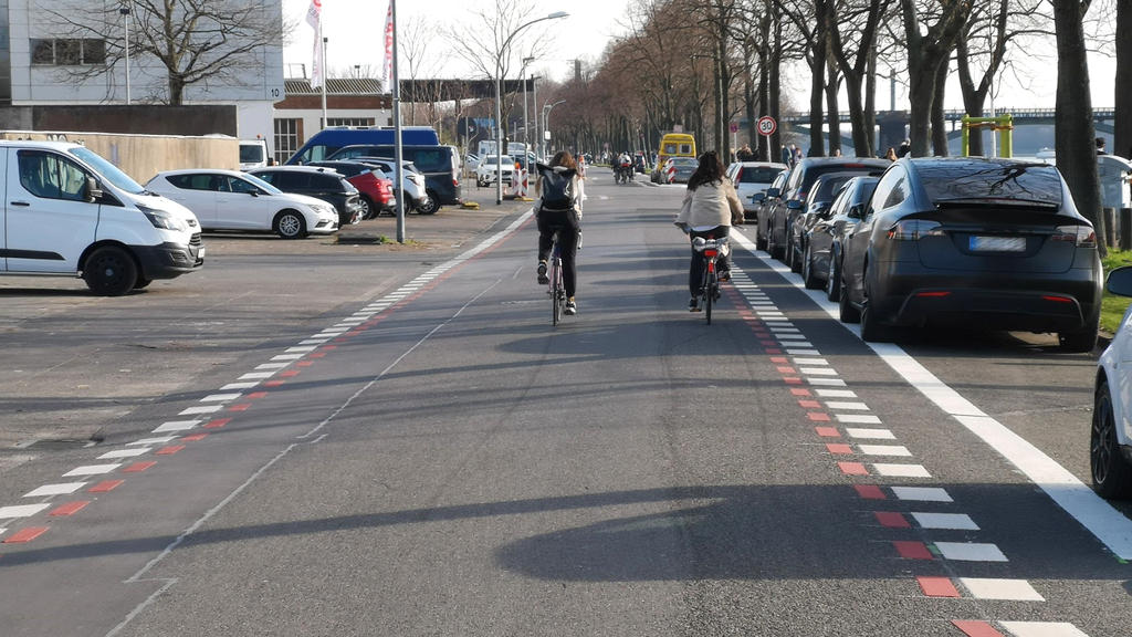 Zwei Radfahrerinnen fahren über die neugestaltete Alfred-Schütte-Allee. Zu sehen sind Parkstände und die neue rote Fahrradstraßenlinie.