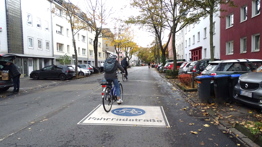 Radfahrende fahren die neue eingerichtete Fahrradstraße Weyertal entlang. Auf dem Boden ist ein großes Fahrradstraßen-Piktogramm zu sehen.