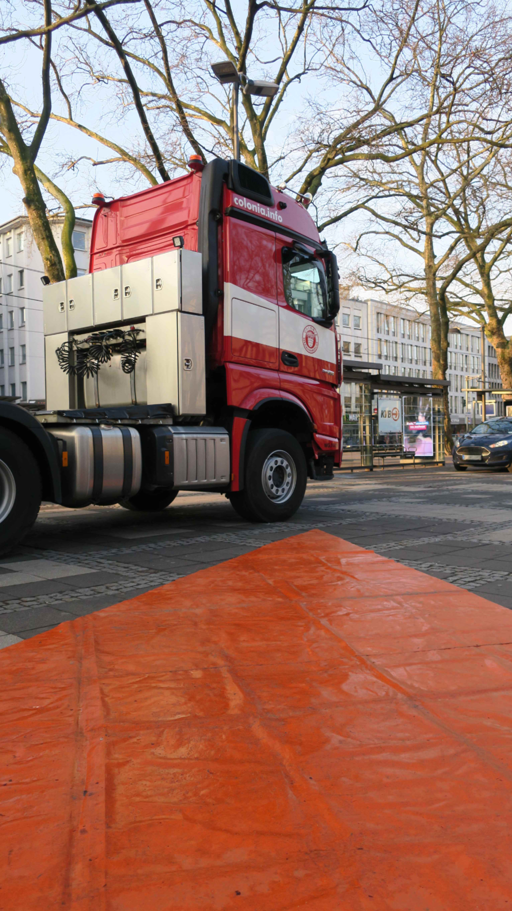 Foto eines großen Lkws. Rechts von diesem in Form eines Dreiecks eine große rote Bodenplane, die den Verlauf des Toten Winkels signalisiert, in dem Menschen von Lkw-Fahrenden schwer gesehen werden können.