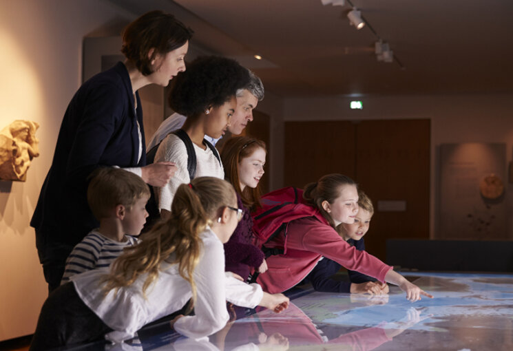 Zwei Erwachsene und mehrere Kinder sind in einem Museum und schauen gemeinsam auf eine Weltkarte, © PantherMedia/Monkeybusiness Images