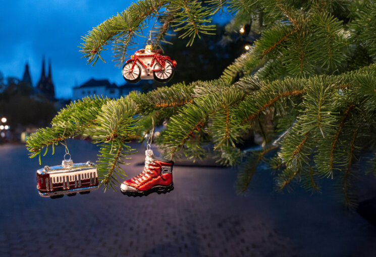 Im Vordergrund sieht man Tannenzweige eines Weihnachtsbaums an denen drei Anhänger in Form einer Bahn, eines Fußes und eines Fahrrades hängen. Im Hintergrund sind die Domspitzen zu sehen., © Stadt Köln
