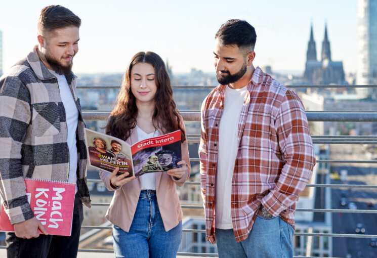 Drei Azubis stehen auf einer Dachterrasse und blättern in der Stadt Intern, im Hintergrund ist der Dom zu sehen., © Rheinisches Bildarchiv/Benita Ruster