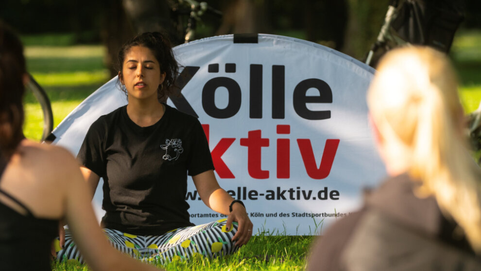 Drei Frauen im Yogasitz auf einer Wiese. Hinter einer Frau ist ein Aufsteller mit der Aufschrift "Kölle aktiv"