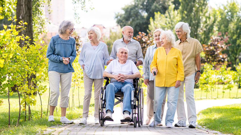 mehrere Senior*innen stehen an einem Frühlingstag nebeneinander in einem Park. In der Mitte sitzt ein Mann im Rollstuhl