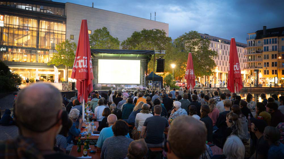 Menschenmenge mit Kopfhörern blicken auf eine Leinwand auf dem Platz  vorm Cinédom im Mediapark.