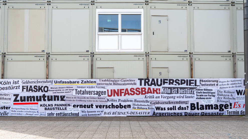 Zu sehen ist ein weißes Banner auf dem die prägnantesten Schlagzeilen der vergangenen 14 Jahre zur Sanierung der Bühnen Köln zu lesen sind. Im Hintergrund stehen Baucontainer. 
