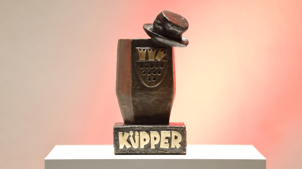 Foto des Karl-Küpper-Preises. Ein Preis aus Metall, auf dem das Kölner Stadtwappen abgebildet ist. Oben rechts auf der Ecke ist ein kleiner Hut auf dem Preis.