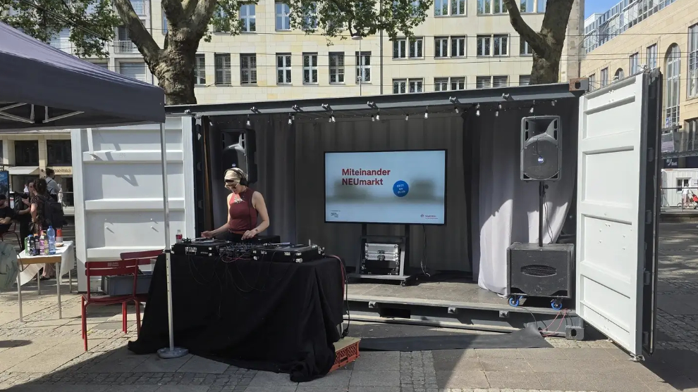 Ein offener Schiffscontainer auf dem Neumarkt in Köln, davor eine DJ an einem Mischpult, interessierte Menschen im Umkreis des Containers.