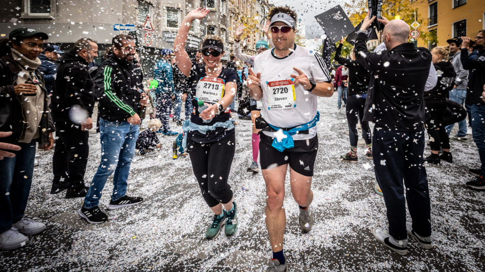 Stimmung an der Strecke des Köln Marathon, im Vordergrund sind zwei Läufer*innen zu sehen, die durch Konfettiregen laufen.