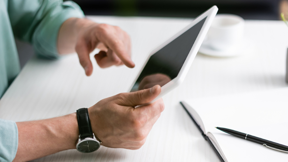 Auf einem Tisch mit weißer Tischdecke liegt ein Hand uny ein Notizbuch. Hände einer nicht weiter zu sehenden Person halten ein Tablet und tippen darauf, alle Bildschirme sind schwarz.