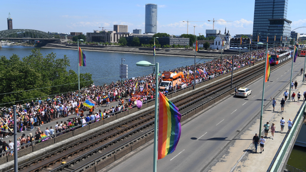 CSD-Parade zieht über die Deutzer Brücke in die linksrheinische Innenstadt. Im Vordergrund weht eine Fahne in Regenbogenfarben.