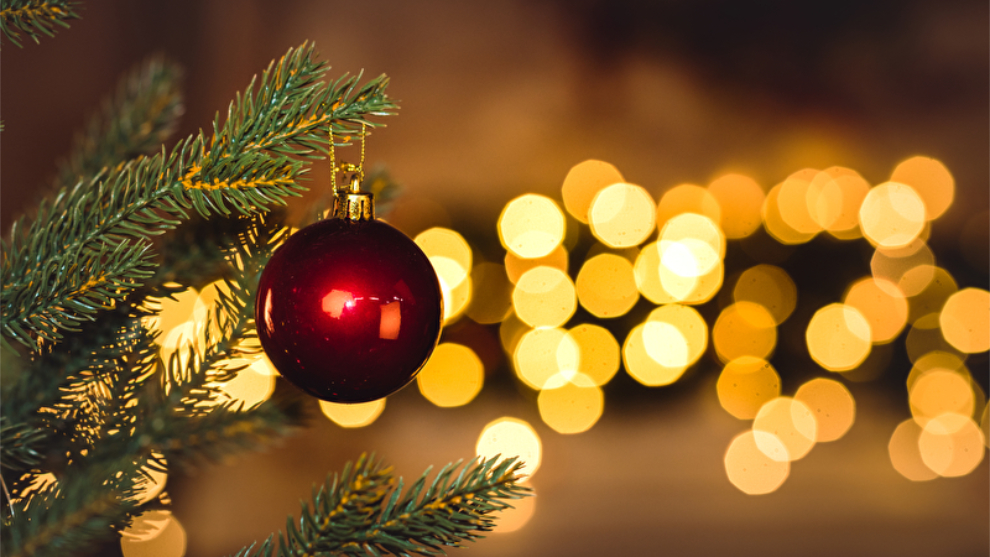 Rote Weihnachtskugel an Weihnachtsbaum. Im Hintergrund liegt eine Lichterkette.