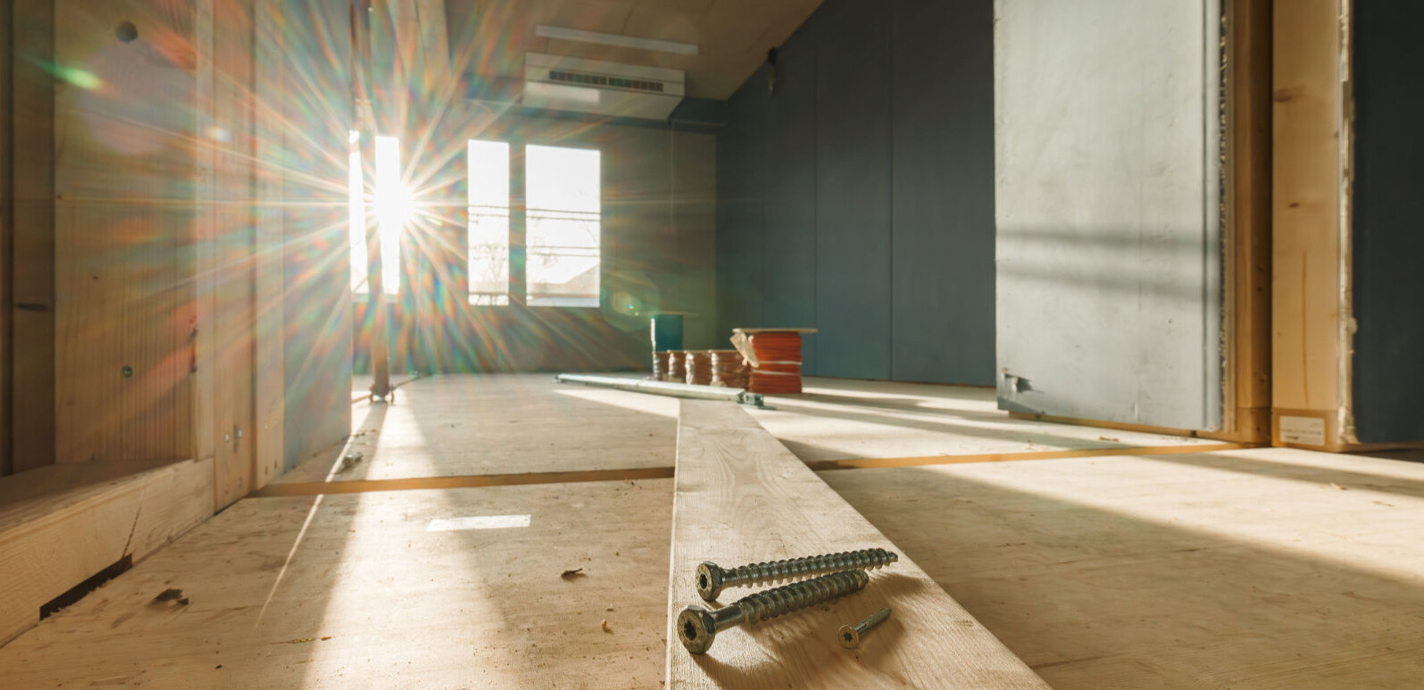 Drei Schrauben auf einer Holzdiele liegend, die in einen künftigen Klassenraum reicht. Durch das Fenster dahinter ist strahl die Sonne herein.