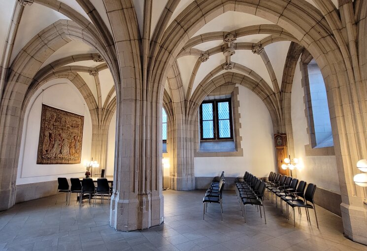 Abgebildet ist ein Trauzimmer mit historischen hohen Rundbögen sowie einem Trautisch mit Stühlen an der linken Seite des Bilds. Weiterhin sind Stühle für Gäste zu sehen, die an der rechten Seite mit Blick zum Hochzeitspaar angeordnet sind. Es handelt sich um die Rentkammer im Historischen Rathaus zu Köln., © Stadt Köln