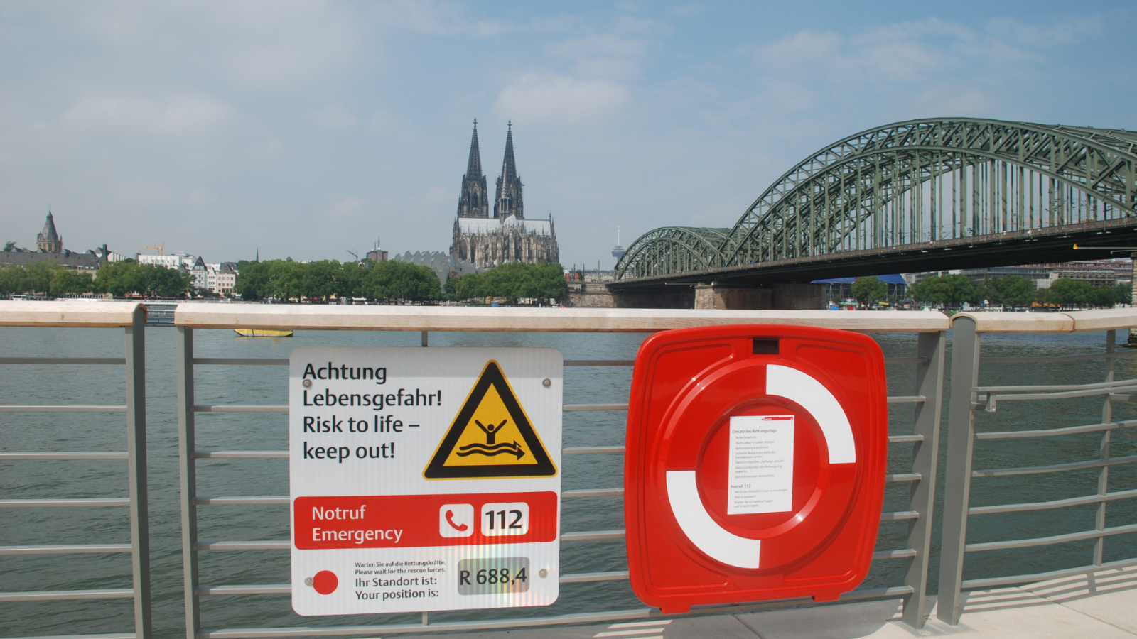 Hinweisschild Achtung Lebensgefahr mit Notrufnummer 112 am Deutzer Rheinufer. Im Hintergrund ist der Kölner Dom und die Hohenzollernbrücke zu sehen.